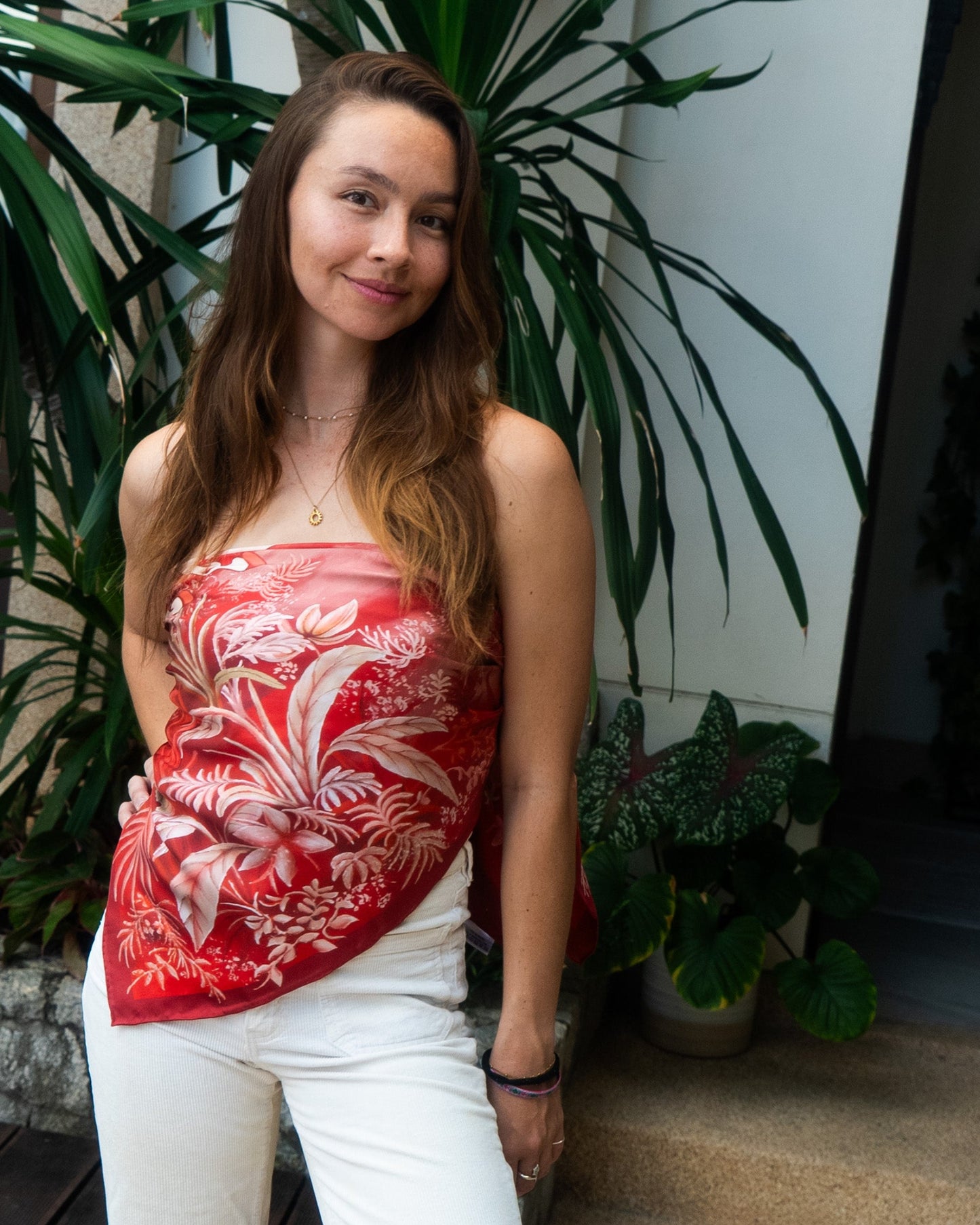 Woman wearing a red botani bloom scarf as a top and white pants standing in front of plants.