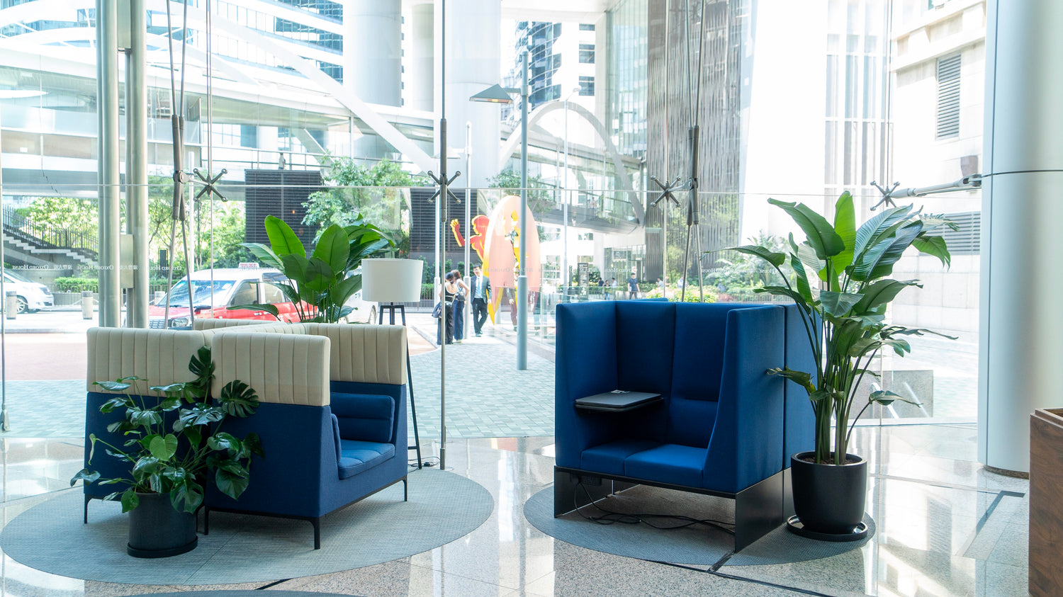Modern lobby with blue and beige seating, potted plants, and city view through large windows.