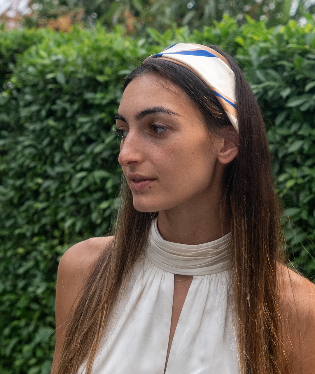 Woman wearing a white botani bloom scarf as a headband with blue accents outdoors against a green hedge.