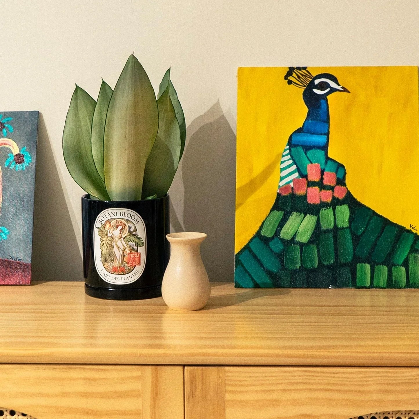 Decorative setup with a potted plant, vase, and painting of a peacock on a wooden surface.