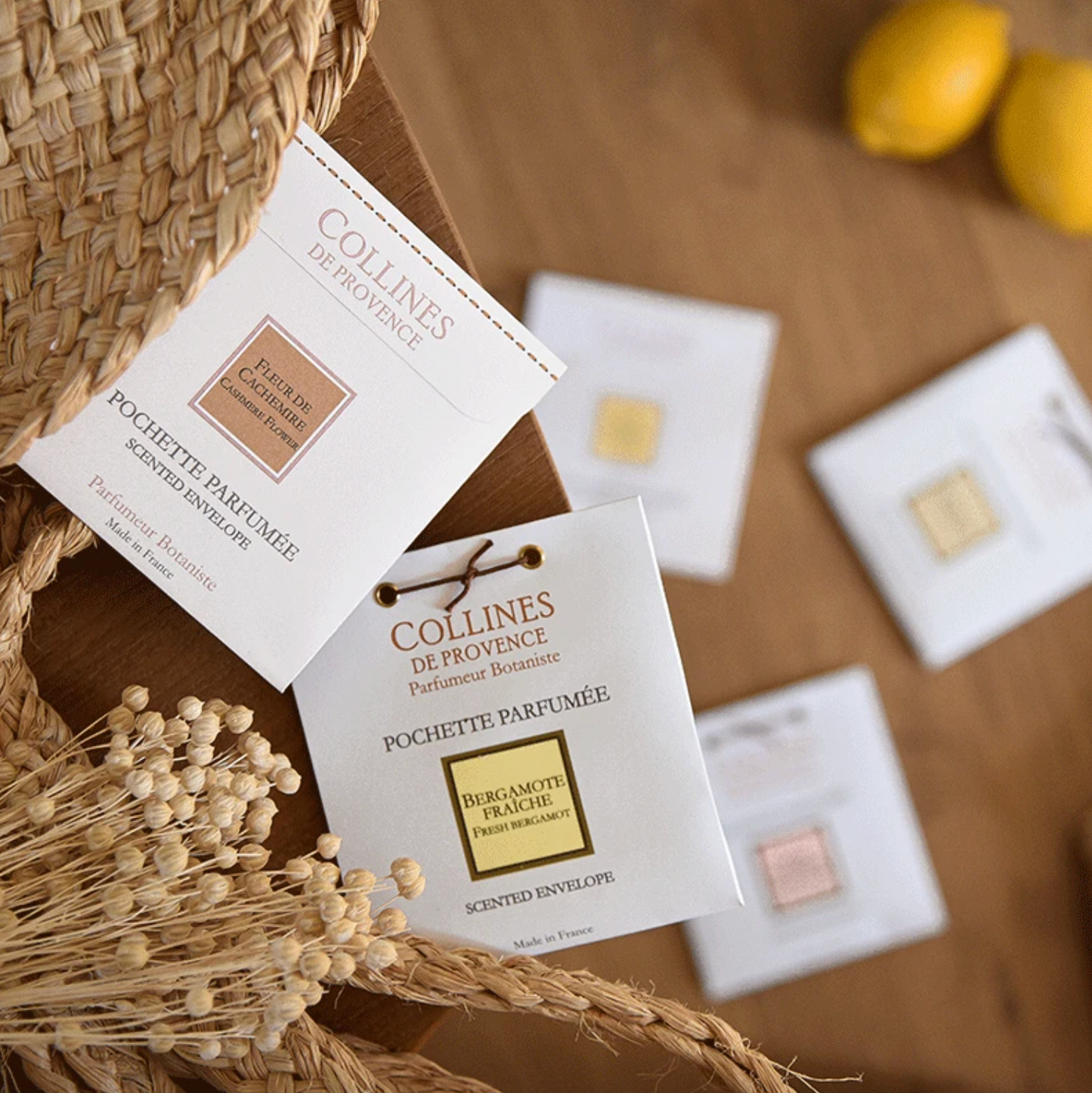 Different Collines de Provence scented envelopes on a wooden surface with a woven basket and lemons.