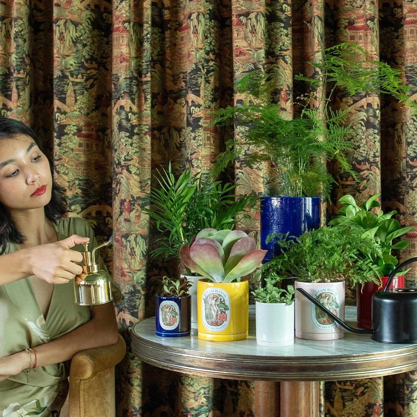 Woman holding a plant mister in front of a table with potted plants against patterned curtains.