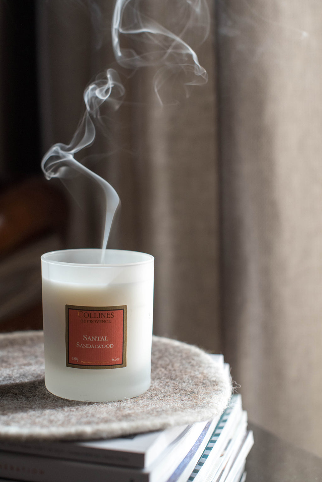 Collines de Provence Sandalwood Scented Candle with smoke rising from it on a textured surface with a blurred background