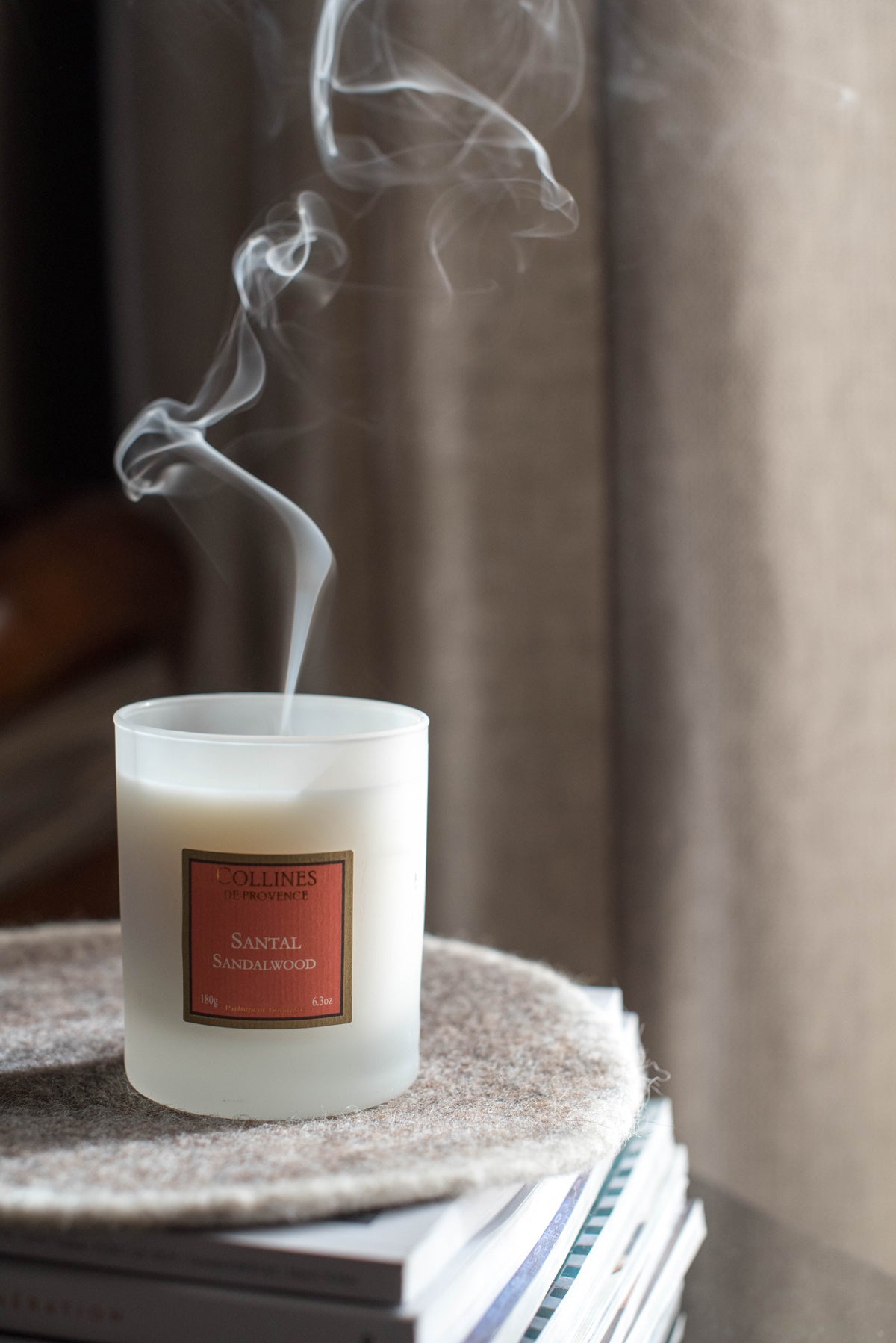 Collines de Provence Sandalwood Scented Candle with smoke rising from it on a textured surface with a blurred background