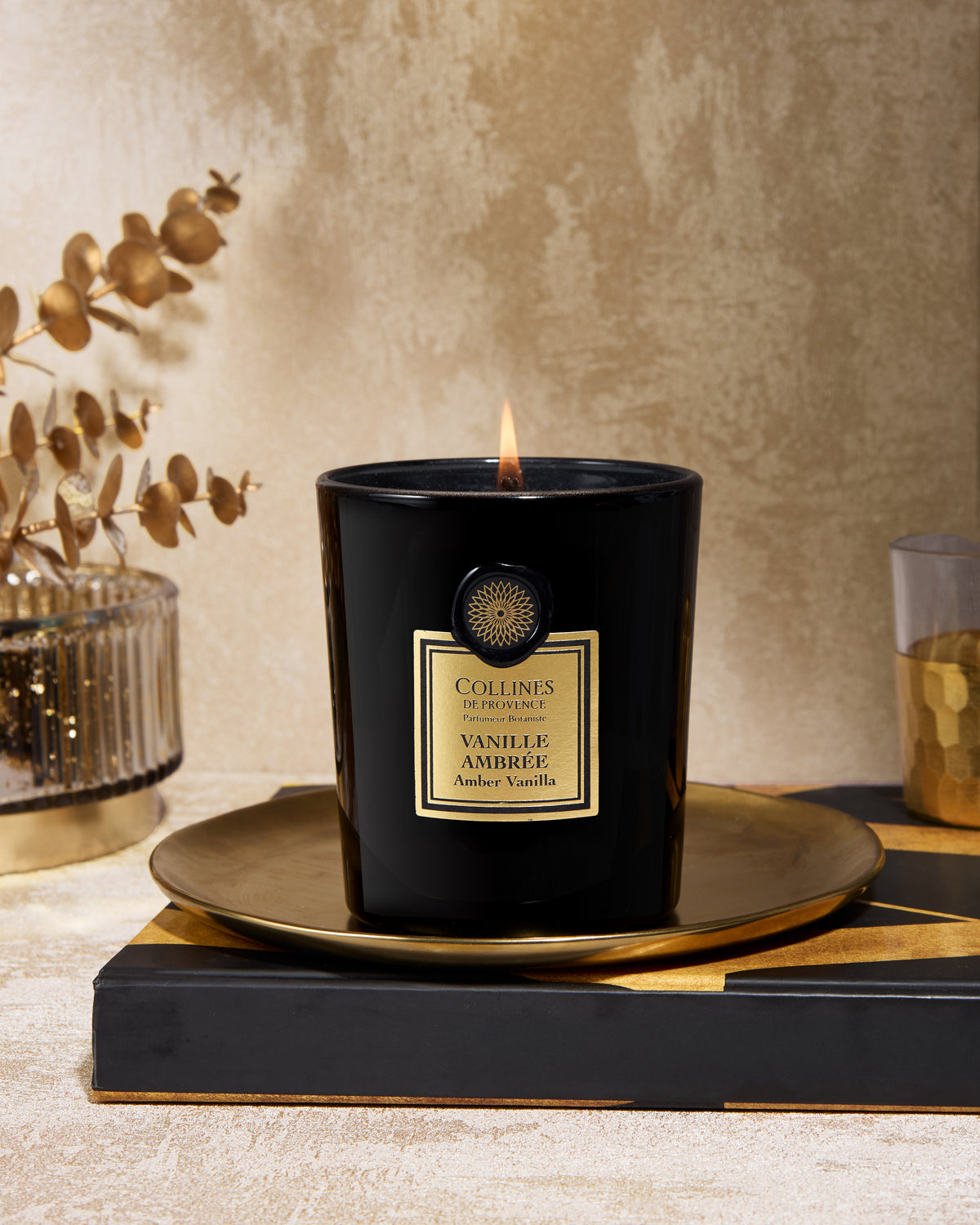 Black scented candle labeled 'Vanille Ambree' or 'vanilla amber' on a decorative stand with a textured beige wall in the background.