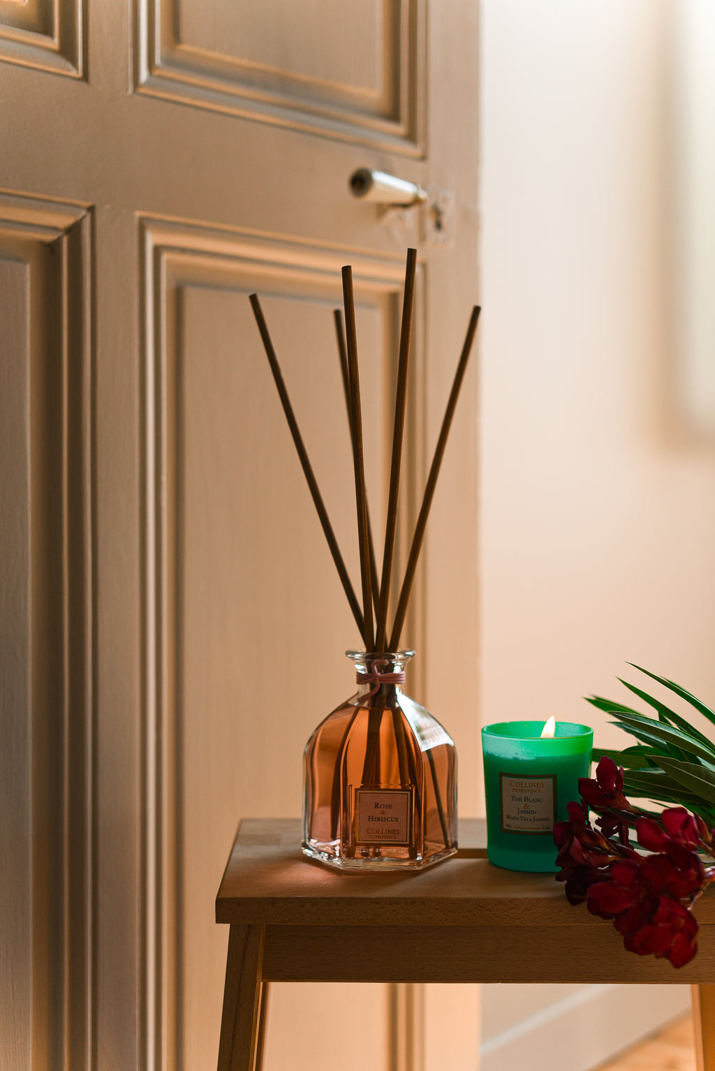 rose and hibiscus diffuser with reeds and a scented candle on a wooden surface against a beige wall.