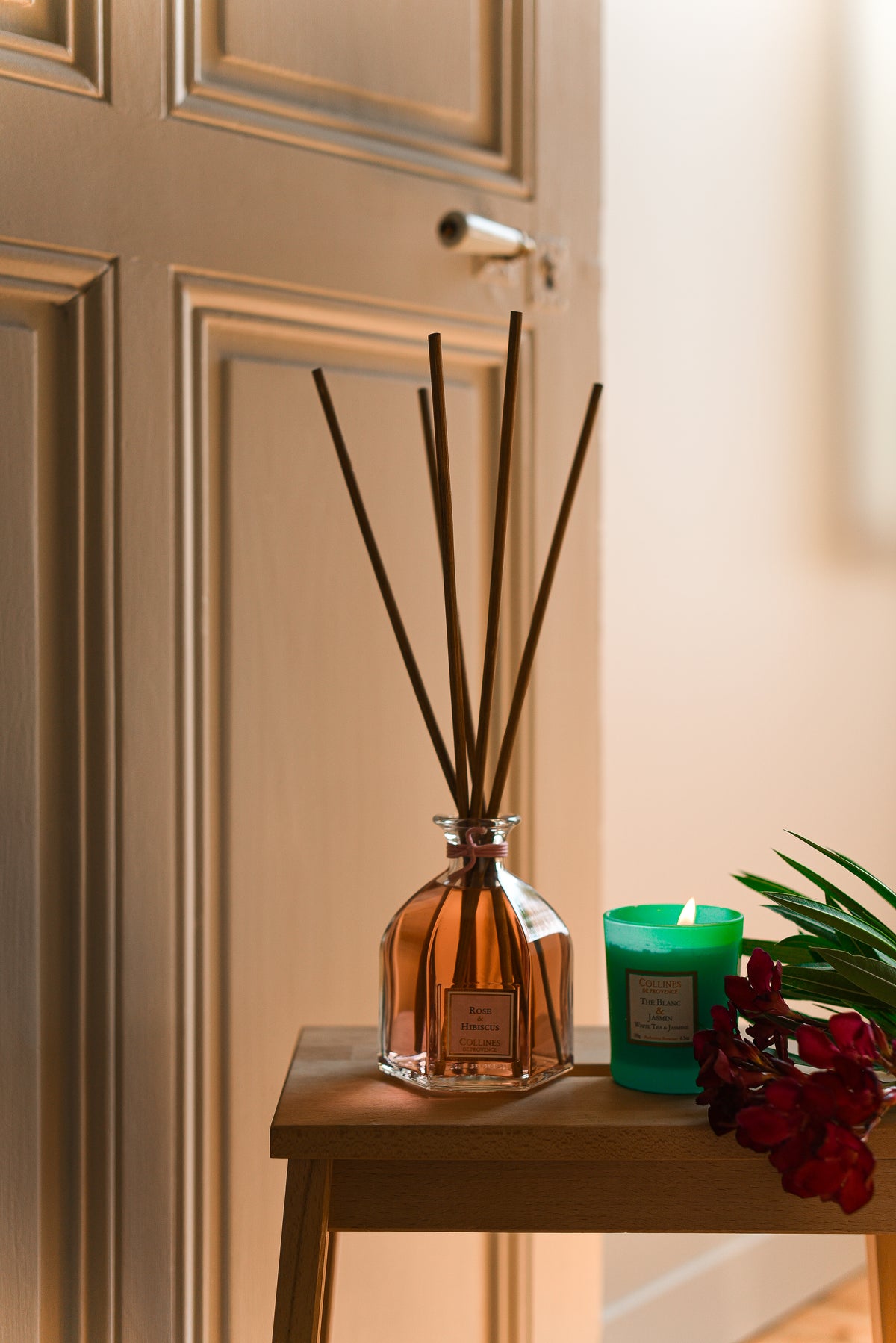 rose and hibiscus diffuser with reeds and a scented candle on a wooden surface against a beige wall.