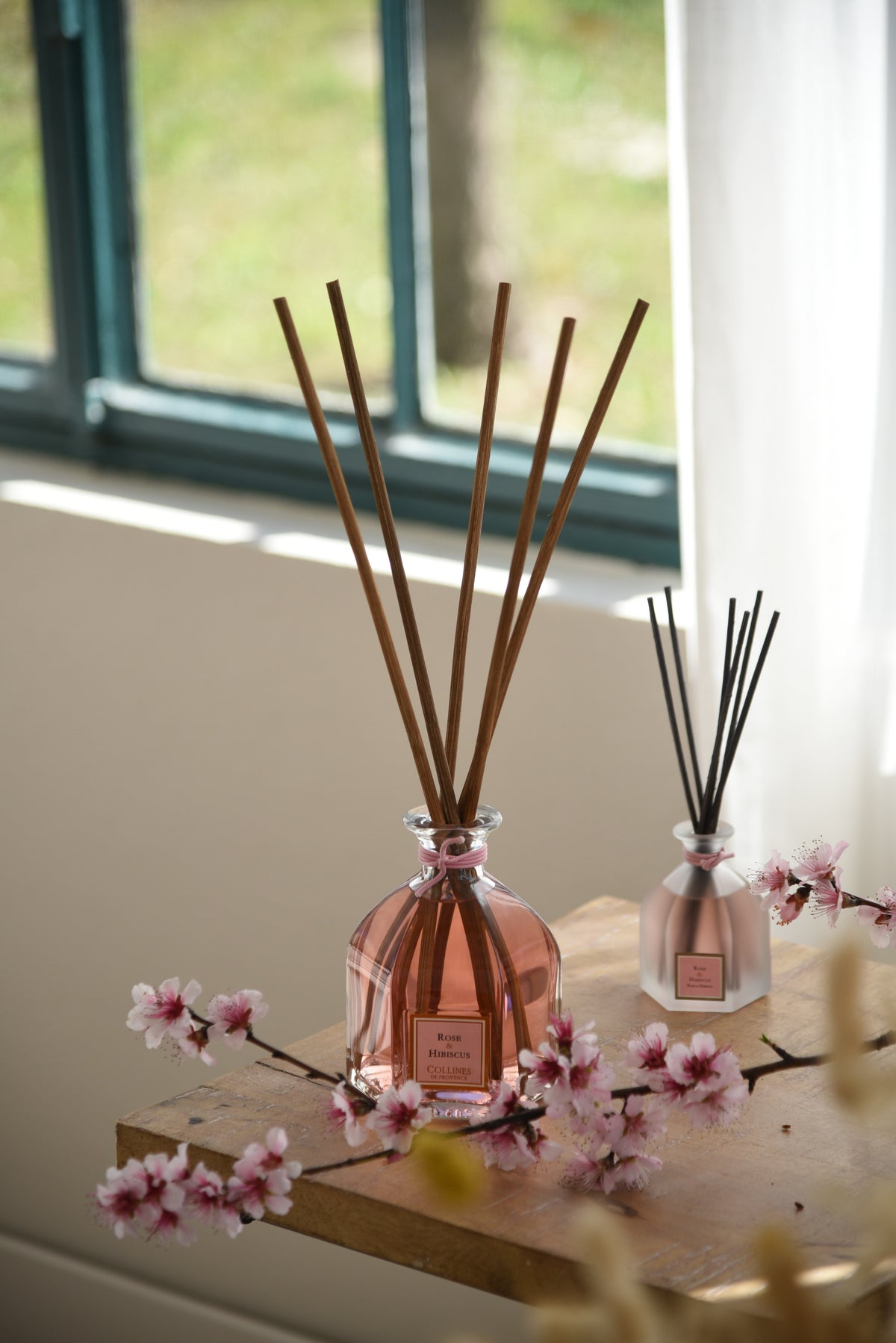 Two rose and hibiscus reed diffusers, one regular size and one large 500ml size, with wooden sticks and pink flowers on a wooden surface.