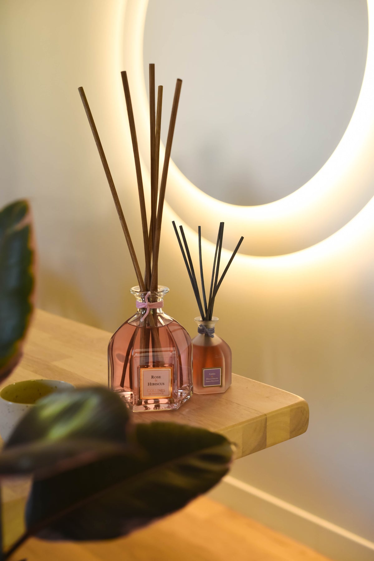 Two diffuser bottles with reeds on a wooden shelf against a softly lit wall.