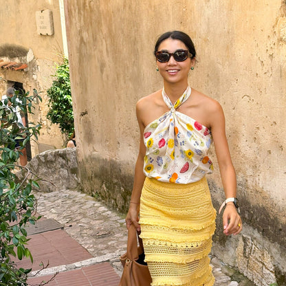 Woman in a floral silk scarf top and yellow skirt standing in front of a stone wall.