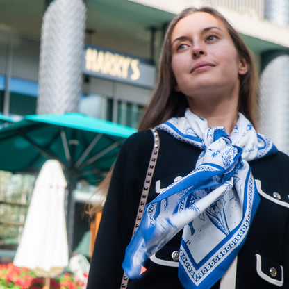 Woman wearing a blue and white botani bloom mistral silk scarf in front of a store with a visible sign.