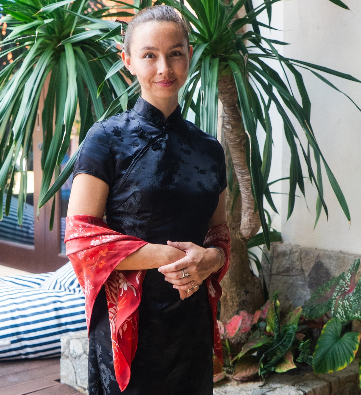 Woman in a black top with silk red scarf standing outdoors with plants in the background