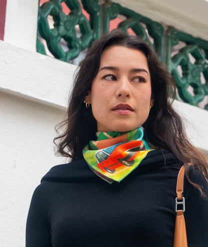 Woman wearing a colorful botani bloom scarf with a car design, standing against a decorative wall.
