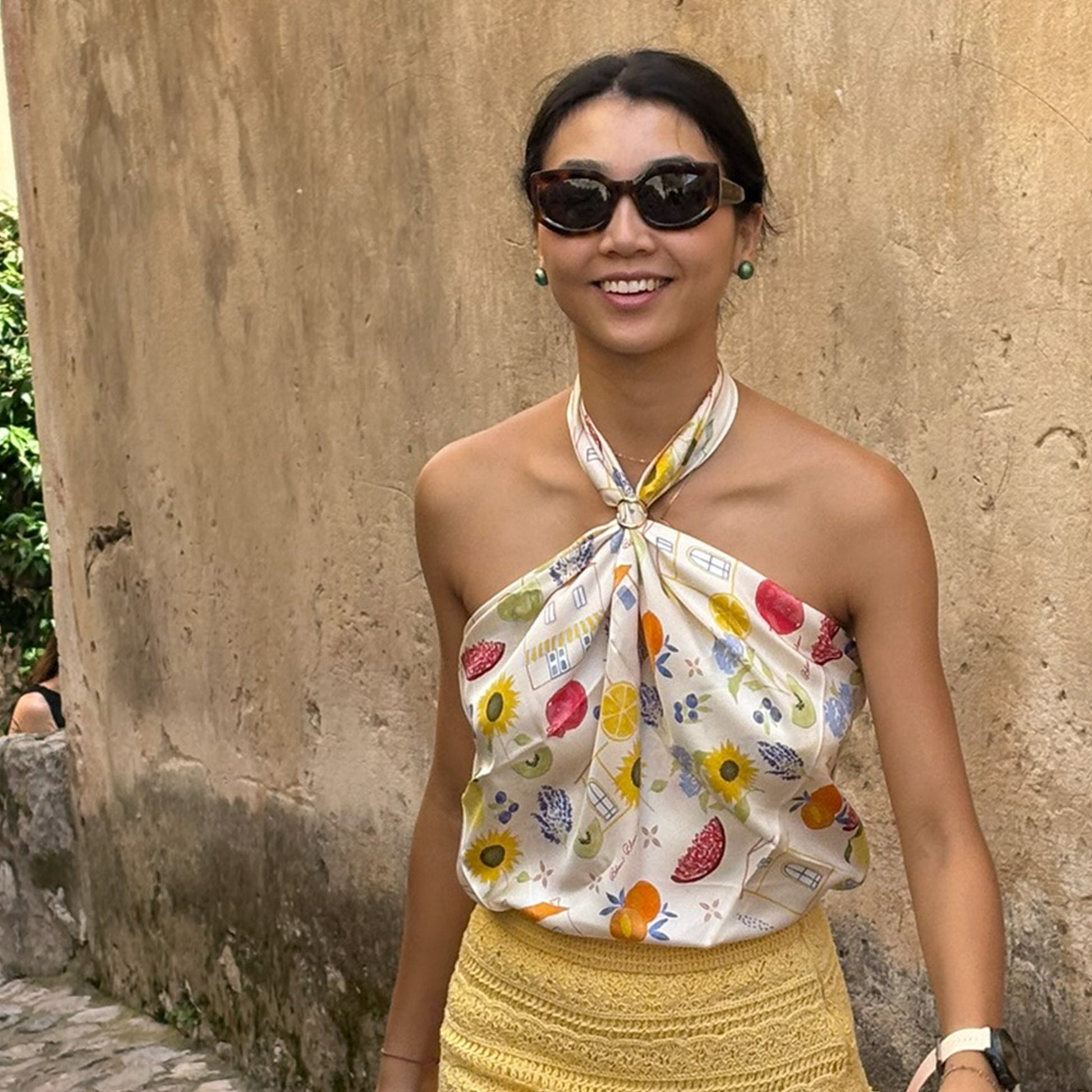 Woman wearing a colorful botani bloom Fruite scarf as a top and yellow skirt against a stone wall.