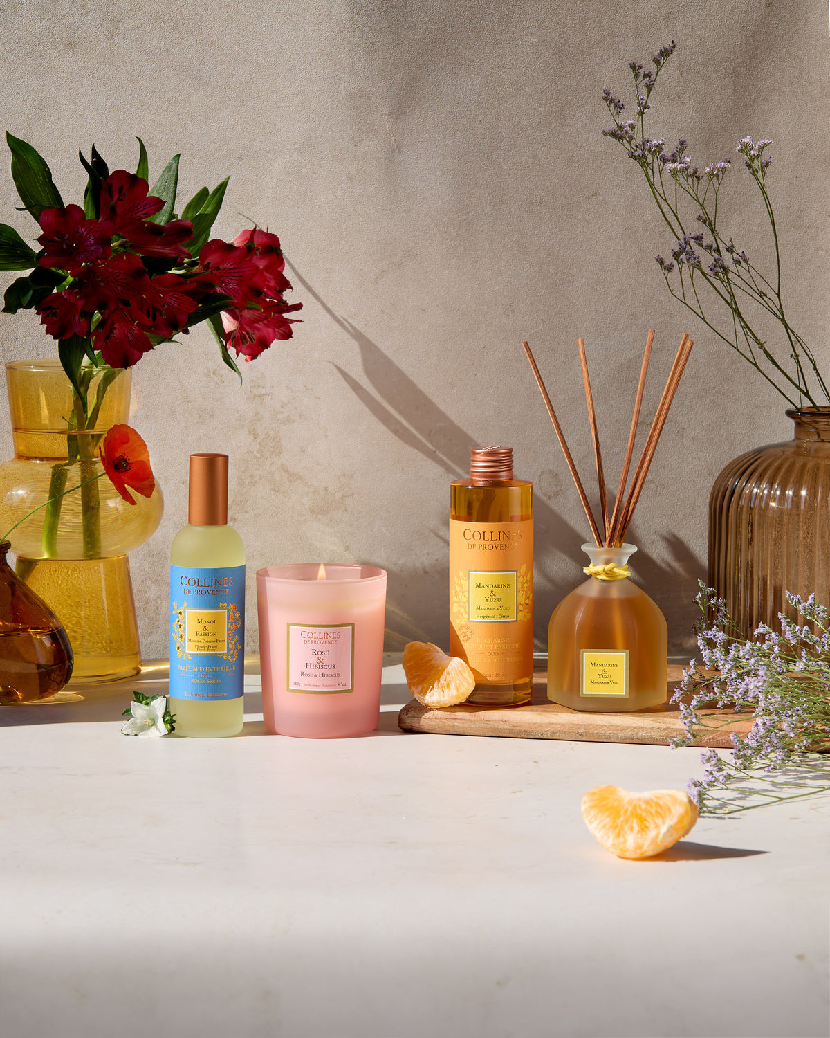 Assorted Collines De Provence products including candle, room spray, reed diffuser, and diffuser refill on a neutral background with flowers and fruits.
